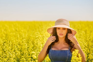 Güneşli bir günde çiçek açan kolza tarlasında poz veren şapkalı güzel bir kadın.