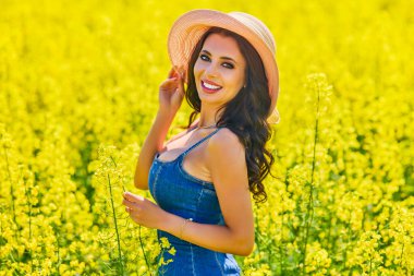Güneşli bir günde çiçek açan kolza tarlasında poz veren şapkalı güzel bir kadın.