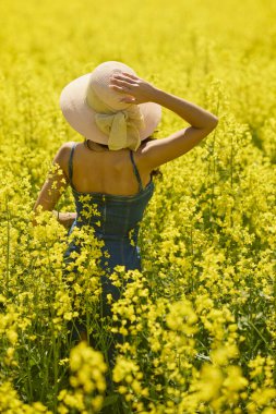 Güneşli bir günde çiçek açan kolza tarlasında poz veren şapkalı güzel bir kadın.