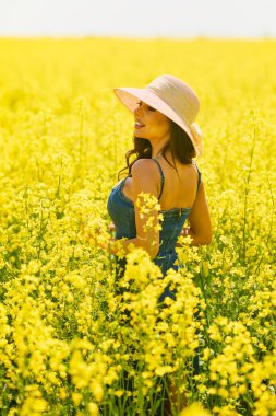 Güneşli bir günde çiçek açan kolza tarlasında poz veren şapkalı güzel bir kadın.