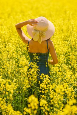 Güneşli bir günde çiçek açan kolza tarlasında poz veren şapkalı güzel bir kadın.