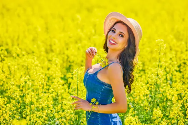 Güneşli bir günde çiçek açan kolza tarlasında poz veren şapkalı güzel bir kadın.
