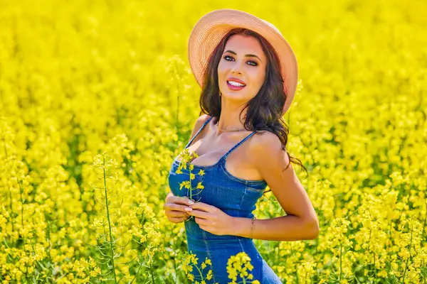 Güneşli bir günde çiçek açan kolza tarlasında poz veren şapkalı güzel bir kadın.