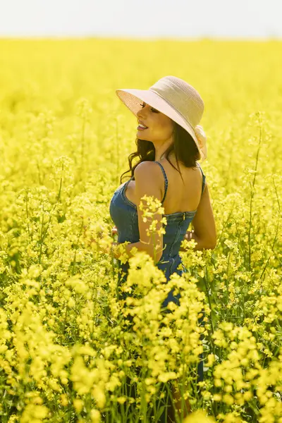 Güneşli bir günde çiçek açan kolza tarlasında poz veren şapkalı güzel bir kadın.