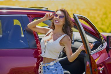 Uzun saçlı güzel bir kadın tarlada kırmızı bir cipin yanında duruyor..