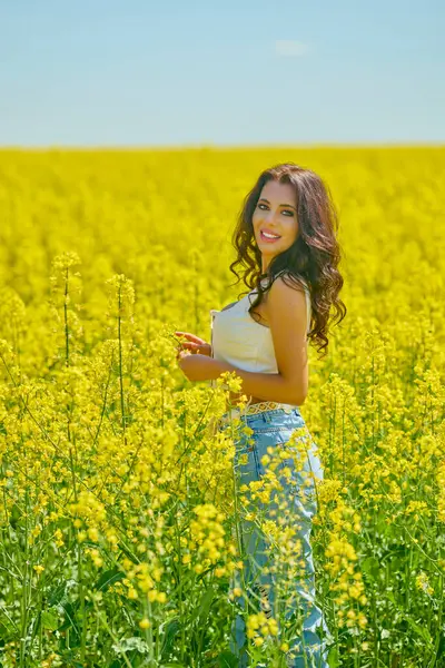 Bluz giymiş uzun saçlı güzel bir kadının portresi ve çiçek açan kolza tohumu tarlasında kot pantolon.