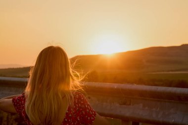 Arkadan gün batımına bakarken görülen bir kadının portresi.