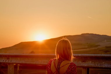 Arkadan gün batımına bakarken görülen bir kadının portresi.