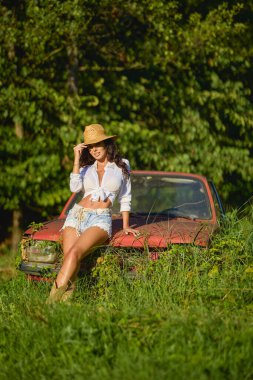 Hasır şapkalı güzel bir kadın bitkilerle çevrili terk edilmiş eski bir arabanın yanında poz veriyor..