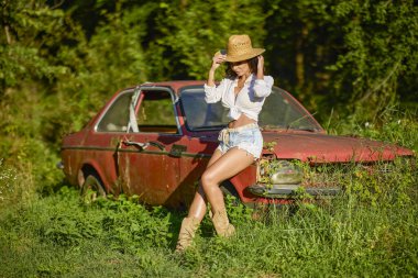 Hasır şapkalı güzel bir kadın bitkilerle çevrili terk edilmiş eski bir arabanın yanında poz veriyor..