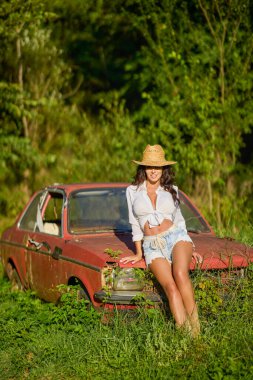 Hasır şapkalı güzel bir kadın bitkilerle çevrili terk edilmiş eski bir arabanın yanında poz veriyor..