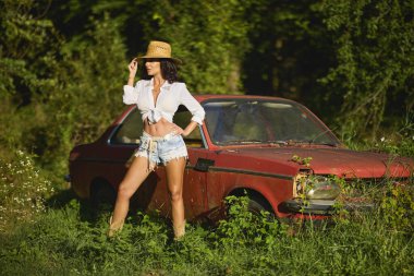 Hasır şapkalı güzel bir kadın bitkilerle çevrili terk edilmiş eski bir arabanın yanında poz veriyor..