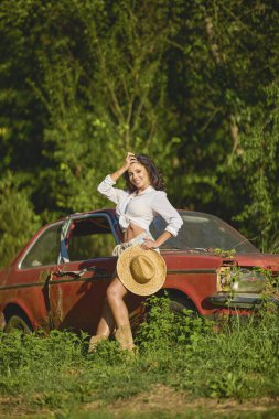 Hasır şapkalı güzel bir kadın bitkilerle çevrili terk edilmiş eski bir arabanın yanında poz veriyor..