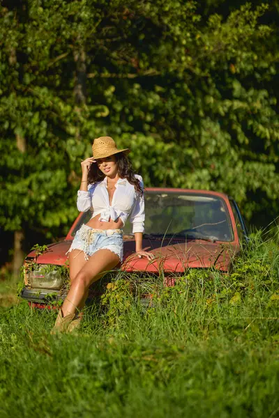 Hasır şapkalı güzel bir kadın bitkilerle çevrili terk edilmiş eski bir arabanın yanında poz veriyor..
