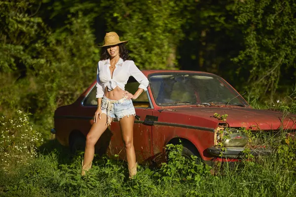 Hasır şapkalı güzel bir kadın bitkilerle çevrili terk edilmiş eski bir arabanın yanında poz veriyor..