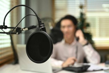 Ev stüdyosunun içinde, erkek radyo sunucusunun arka planda oturduğu yoğunlaştırıcı bir mikrofonun görüntüsünü kapat..