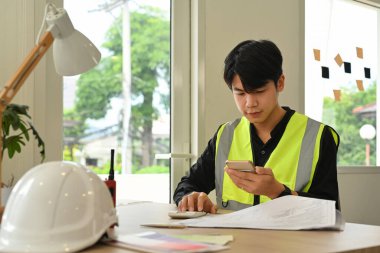 Genç mimar cep telefonu kullanıyor, işyerinde bir inşaat planı üzerinde çalışıyor..