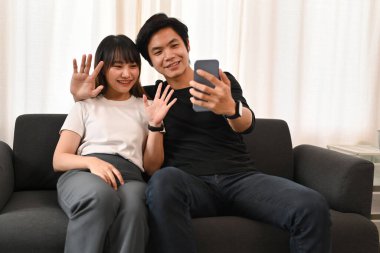 Mutlu genç çift selfie çekiyor, akıllı telefondan video çekiyor ve evde kanepede oturuyor..