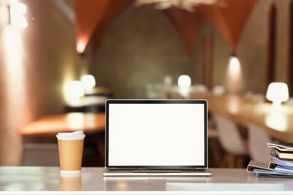 Laptop on white table with blurred restaurant in background. Blank screen for your product display or design montage.