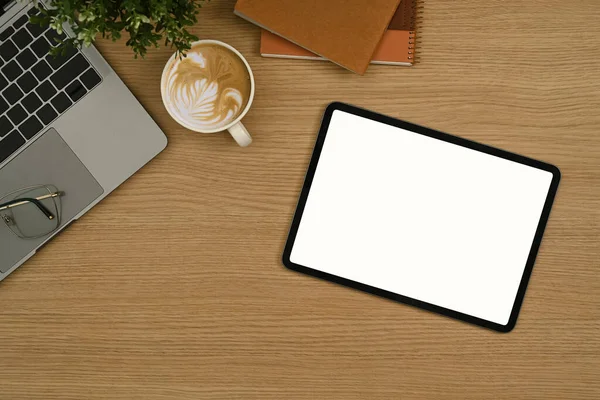Flat lay digital tablet with empty display, coffee cup, laptop and notebook on wooden office desk.