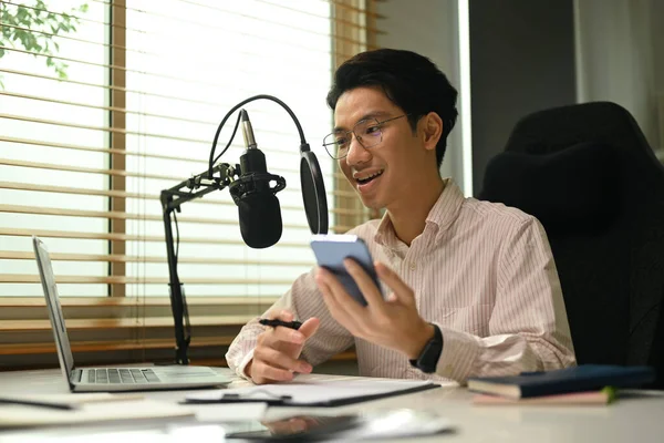 Asian man radio host using smart phone and talking through microphone to recording podcast in small home studio.