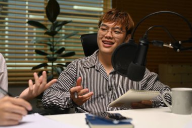 Gülümseyen erkek radyo sunucusu stüdyoda ses yayını yaparken misafirine bakıyor ve onunla konuşuyor..