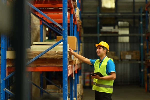 Asian male manager wearing safety hardhat and vest checking quantity of storage product on shelf in a large retail warehouse.