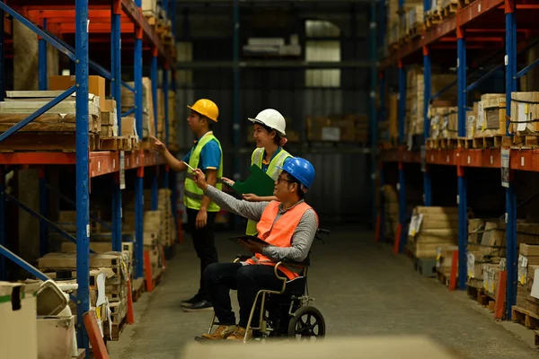 Middle aged male manager in wheelchair and young workers inspecting stock product in a large warehouse.