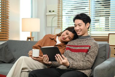 İnternetten dijital tabletle alışveriş yapan neşeli genç çift evdeki kanepede dinleniyor. İnsanlar ve teknoloji kavramı.