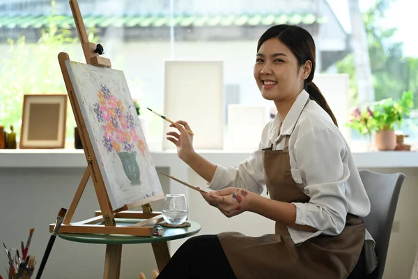 Smiling creative woman painting picture on canvas with oil paints in bright home studio. Leisure activity and art concept.