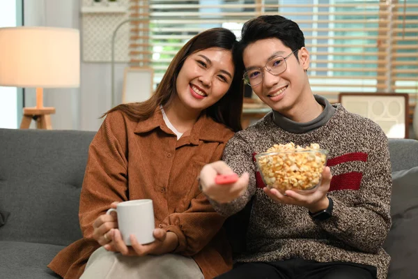 Happy young asian couple eating popcorn and watching movie at home. Entertainment and leisure activity concept.