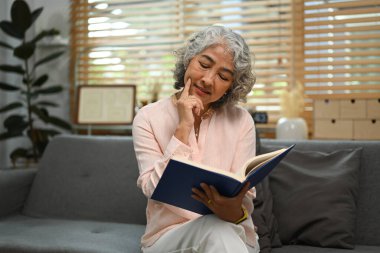 Oturma odasındaki rahat koltukta oturan, bir fincan kahve içip kitap okuyan rahat yaşlı bir kadın..