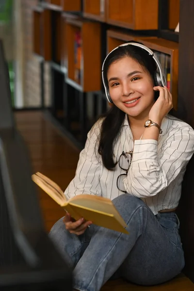 ヘッドフォンで音楽を聴く陽気なアジアの女性学生と大学図書館で本を読む — ストック写真