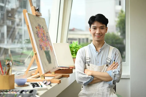 Portrait of handsome male artist standing with arms crossed front of easel in bright art studio and smiling to camera.