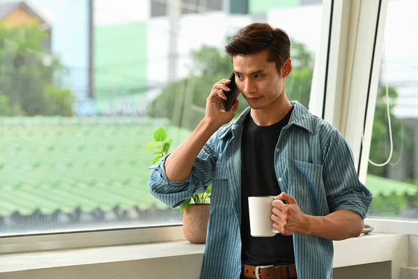 Jóképű Fiatalember Kezében Kávéscsésze Beszél Mobiltelefon Miközben Áll Irodai Ablak — Stock Fotó