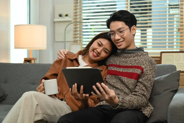 居心地の良いリビングルームでリラックスしながら デジタルタブレットでビデオやブラウジングインターネットを見て愛するアジアのカップルのイメージ — ストック写真
