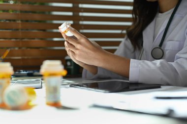 Elinde ilaç şişeleri olan beyaz önlüklü bir doktorun tıbbi ofisinde çalışırken çekilmiş bir fotoğrafı..