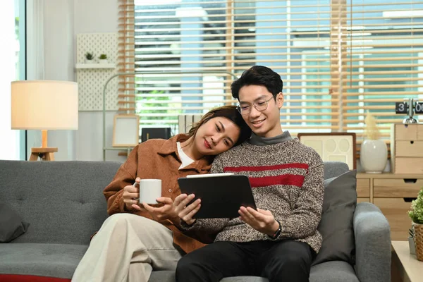 若いアジアのカップルを愛しビデオを見たり デジタルタブレットでインターネットを閲覧しながら 居心地の良いリビングルームに座って — ストック写真