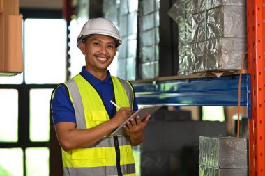 Karton kutuların yanında dikilirken dijital tablet kullanarak güvenlik yeleği giyen yakışıklı bir depo çalışanı..