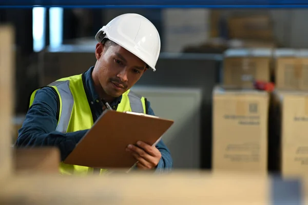 Asian male manager wearing hardhat and reflective jacket checking inventory in a warehouse with shelves full of cardboard boxes.