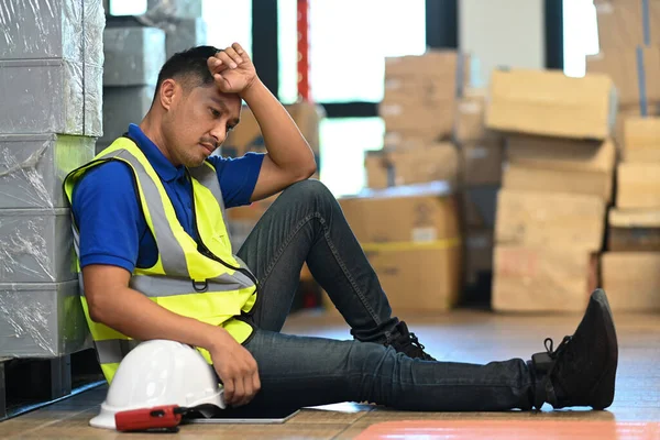 Tired male warehouse worker sitting on floor and taking break from hard work in large retail warehouse.