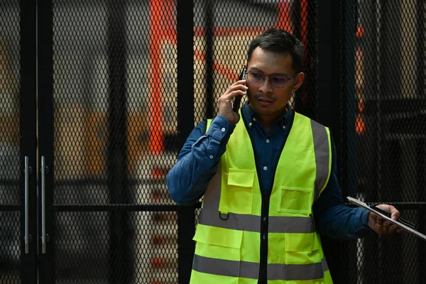 Image of serious male manager in yellow vest talking on mobile phone while standing in the retail warehouse.