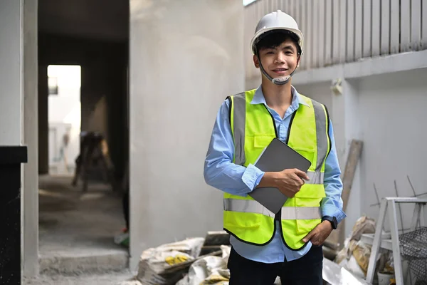 Portrait of architect man in safety helmet and reflective vest standing front of construction site and smiling to camera.