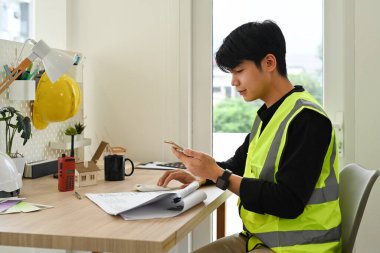 Akıllı telefon kullanan mühendisin yan görüntüsü modern ofisindeki inşaat planlarını inceliyor..