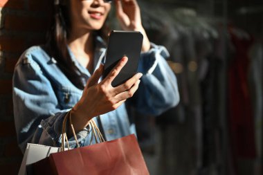 Şık bir kadın alışveriş torbaları taşıyor ve güneşli bir günde açık havada akıllı telefon kullanıyor..