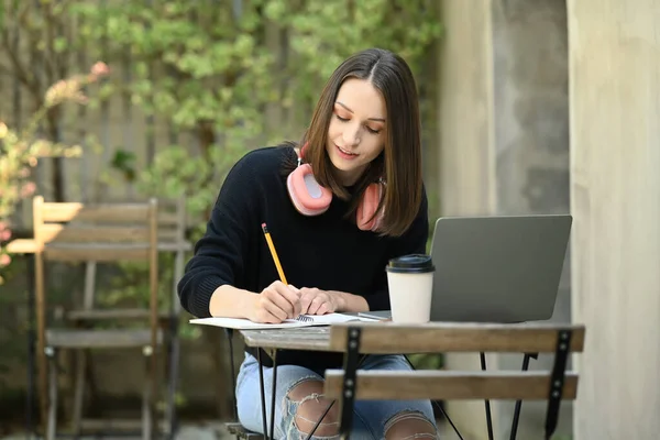 stock image Pretty caucasian young woman writing down in notebook and using laptop while sitting at outdoor.