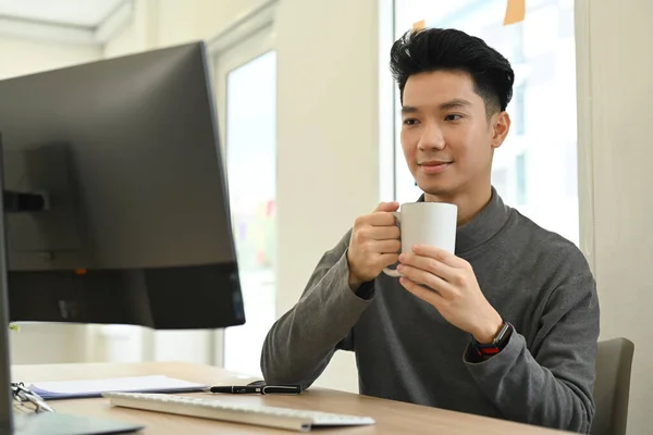 Smiling asian man holding cup of coffee and watching online webinar on computer screen.