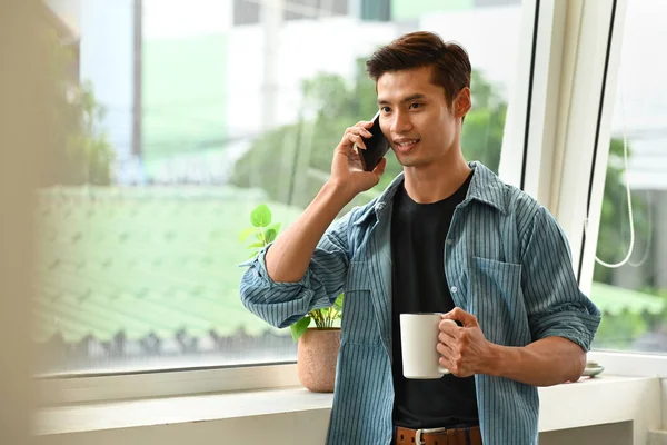 Handsome asian man holding coffee cup and talking on mobile phone while standing near the office window.