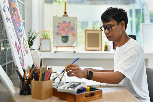 Asian man artist sitting in art studio and painting picture on canvas with oil paints. Art and leisure activity concept.