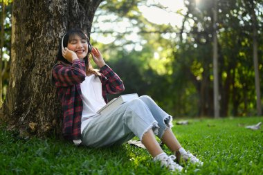 Rahatlamış genç bir kadın, güneşli bir günde yeşil çimlerin üzerinde otururken kulaklıkla müzik dinlemekten zevk alır..